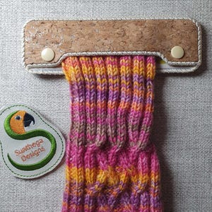 May include: A cork and fabric sock holder with a white button on each side. The holder is holding a colorful knitted sock with pink, orange, yellow, and brown stripes.
