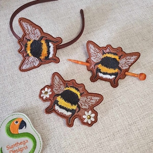 May include: Three embroidered bumble bee hair accessories. One bee is on a brown headband, one bee is on a wooden hair stick, and one bee is on a brown leather patch with white flowers.