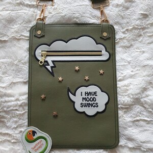 May include: An olive green crossbody bag with a cloud and lightning bolt design. It has a gold zip, star accents, and the phrase "I HAVE MOOD SWINGS." Includes a small, round patch with a parrot illustration and the text "Sunthega Designs."