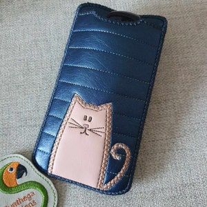 May include: A blue quilted phone case with a pink cat design. The cat has a white face with black whiskers and eyes.