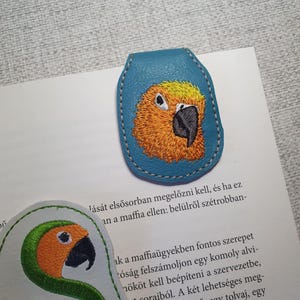 May include: A teal leather bookmark with a detailed embroidered image of a yellow and orange parrot. The bookmark is stitched with white thread. A second bookmark with a parrot design is also visible.