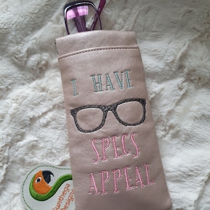 May include: A pink leather glasses case with the text "I have specs appeal" embroidered on it. The case has a black and white pair of glasses embroidered on it. A green and orange parrot logo is in the lower left corner.