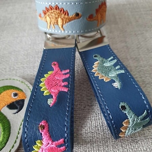 Può includere: Due portachiavi in ​​cuoio blu con dinosauri ricamati. Un portachiavi ha dinosauri rosa e l'altro ha dinosauri verdi. I portachiavi sono attaccati a anelli portachiavi d'argento.
