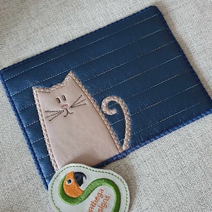 May include: A blue quilted fabric coaster with a beige embroidered cat design. The coaster has a green and orange embroidered patch that says "Swathega Designs".