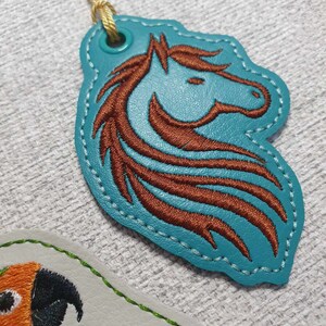 May include: A teal blue leather key chain with a brown embroidered horse head design. The key chain has a gold cord and a metal ring.