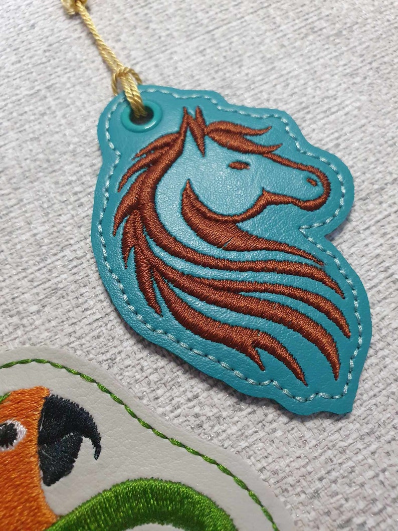 May include: A teal blue leather key chain with a brown embroidered horse head design. The key chain has a gold cord and a metal ring.