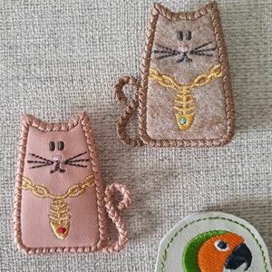 May include: Two felt cat ornaments with gold chains and jeweled pendants. The cats are brown and pink with embroidered details. A green and orange parrot ornament is in the background.