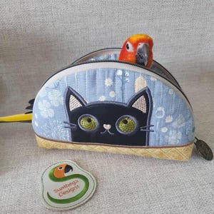 May include: A blue and white fabric pouch with a black cat embroidered on the front. The pouch has a zipper closure and a yellow and orange parrot peeking out of the top. The pouch has a label that says "Sunthaga Designs".