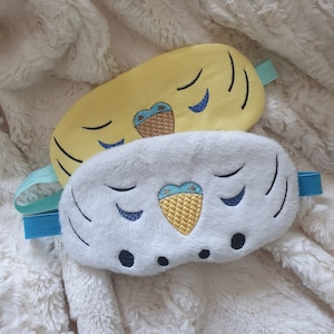 May include: Two eye masks, one yellow with a blue and white embroidered design and one white with a blue and white embroidered design. Both masks have a blue ribbon.