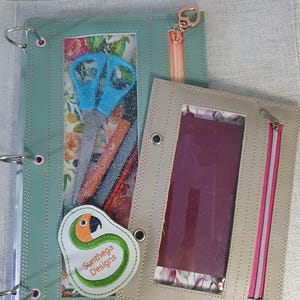 May include: A sage green three-ring binder with a clear window, a peach zipper, and a parrot patch labeled Sunthega Designs, paired with a smaller beige zippered pouch featuring a clear window and a dark purple interior insert.