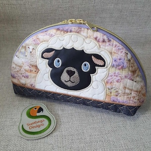 May include: A half-circle shaped zippered pouch with a black sheep face embroidered on the front. The pouch is made of quilted fabric with a floral pattern and a denim trim. The zipper is gold and the pouch is closed. A small embroidered patch with the text "Sunthera Designs" and a parrot is in the lower left corner.