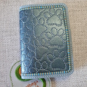 May include: A teal wallet featuring a paw print pattern, with a stitched design and a striped border. The wallet is next to an avocado-shaped patch with the text "Sunthega Designs".