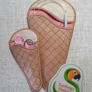 May include: Two brown felt ice cream cone shaped needle holders. The larger cone has a white felt pocket with a clear plastic thread holder and a silver crochet hook. The smaller cone has a pink felt pocket with a pair of small scissors and a pink and white patterned felt circle. A green felt patch with a parrot design and the text "Sunthega Designs" is below the cones.