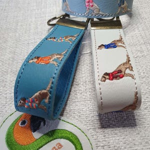 May include: A collection of leather accessories featuring embroidered dog designs. Includes a light blue bracelet and keychains in teal and white. The dogs are depicted wearing colorful sweaters. The accessories have gold-colored hardware.