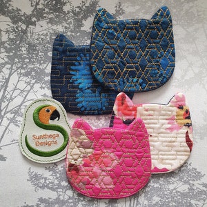 Op de afbeelding: Vier gestikte onderzetters in de vorm van een kat met een geometrisch patroon. De onderzetters zijn in tinten blauw, roze en wit. De onderzetters zijn gemaakt door Sunthaga Designs.
