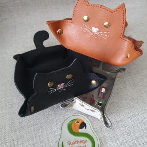 May include: Three cat-shaped trays in black, brown, and white leather. Each tray features embroidered whiskers and a pink nose. Gold rivets secure the tray's edges. A green and white patch with a parrot design and the text "Sunthega Designs" is also visible.