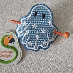 May include: A blue ghost with white embroidered details and black eyes is attached to a wooden hair stick. The hair stick is orange and brown.