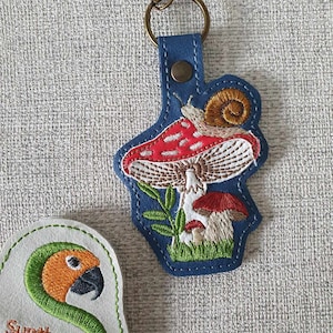 May include: A blue leather keychain with an embroidered mushroom, snail, and leaves. The mushroom is red and white with a brown stem. The snail is brown with a yellow shell. The leaves are green. The keychain has a metal ring and a metal clasp.