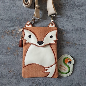 May include: A brown and white fox-shaped crossbody phone bag with a white strap. The bag has a zipper closure and a small tassel. The bag has a green embroidered patch with the text "Sunthaga Designs" and a parrot.