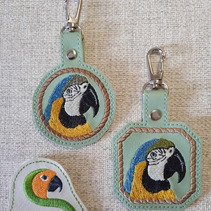 May include: Two green leather keychains with a blue and yellow macaw embroidered on them. The keychains are round and square in shape. A small white leather patch with a green snake and the text "Sunthaga Designs" is also visible.