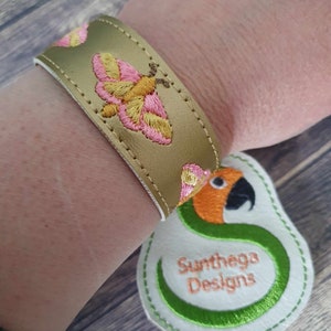 Könnte beinhalten: Ein goldfarbenes Lederarmband mit einem gestickten Schmetterlingsmotiv in Pink und Gelb. Das Armband befindet sich am Handgelenk einer Person. Ein weißes Stoff-Etikett mit einem grünen gestickten Papagei und dem Text "Sunthaga Designs" befindet sich in der unteren rechten Ecke des Bildes.