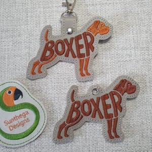 Op de afbeelding: Twee Boxer-hondvormige sleutelhangers en een ronde patch. De sleutelhangers zijn grijs met bruine geborduurde details en het woord "BOXER". Eén sleutelhanger heeft een metalen sluiting. De patch heeft een papegaai-ontwerp en de tekst "Sunthega Designs".
