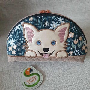 May include: A white dog with blue eyes peeks out from a half-circle zippered pouch with a floral pattern. The pouch is made of quilted fabric and has a brown zipper pull. A green and orange parrot logo is visible below the pouch.