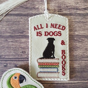 May include: A white leather bookmark with a red embroidered saying "All I need is dogs & books". A black silhouette of a dog sitting on top of a stack of books is embroidered on the bookmark. The bookmark has a white tassel.