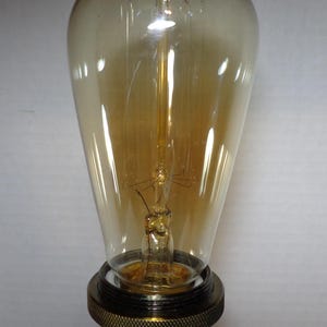 K&ouml;nnte beinhalten: Eine Edison-Gl&uuml;hbirne im Vintage-Stil mit einem tropfenf&ouml;rmigen, bernsteinfarbenen Glasgeh&auml;use. Die Gl&uuml;hbirne hat einen sichtbaren Gl&uuml;hfaden und ist auf einem Messingsockel mit einem strukturierten, schwarzen Ring montiert. Diese dekorative Gl&uuml;hbirne ist f&uuml;r ein warmes, einladendes Leuchten konzipiert.