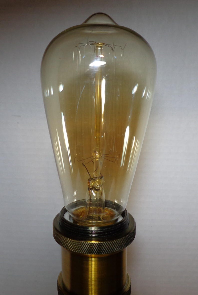K&ouml;nnte beinhalten: Eine Edison-Gl&uuml;hbirne im Vintage-Stil mit einem tropfenf&ouml;rmigen, bernsteinfarbenen Glasgeh&auml;use. Die Gl&uuml;hbirne hat einen sichtbaren Gl&uuml;hfaden und ist auf einem Messingsockel mit einem strukturierten, schwarzen Ring montiert. Diese dekorative Gl&uuml;hbirne ist f&uuml;r ein warmes, einladendes Leuchten konzipiert.