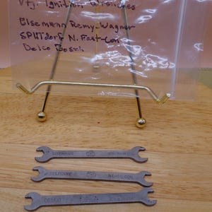 May include: A collection of vintage ignition wrenches, including brands like Eisemann, Splitdorf, and Delco. The wrenches are made of metal and are displayed on a wooden surface. The image also includes a handwritten note.