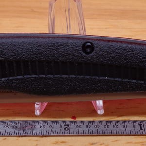 May include: A black folding knife with a textured handle and a silver blade release. The knife is shown closed, with a ruler below it. The handle has a ridged design and a silver screw.