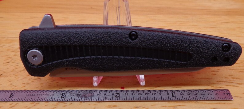 May include: A black folding knife with a textured handle and a silver blade release. The knife is shown closed, with a ruler below it. The handle has a ridged design and a silver screw.