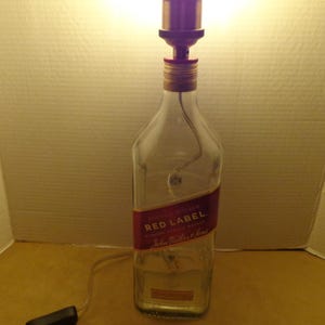 K&ouml;nnte beinhalten: Eine Lampe aus einer recycelten Glasflasche mit einem roten Etikett "John Walker & Sons Red Label". Sie hat eine kupferfarbene Lampenfassung und einen schwarzen Ein-/Ausschalter am Kabel.