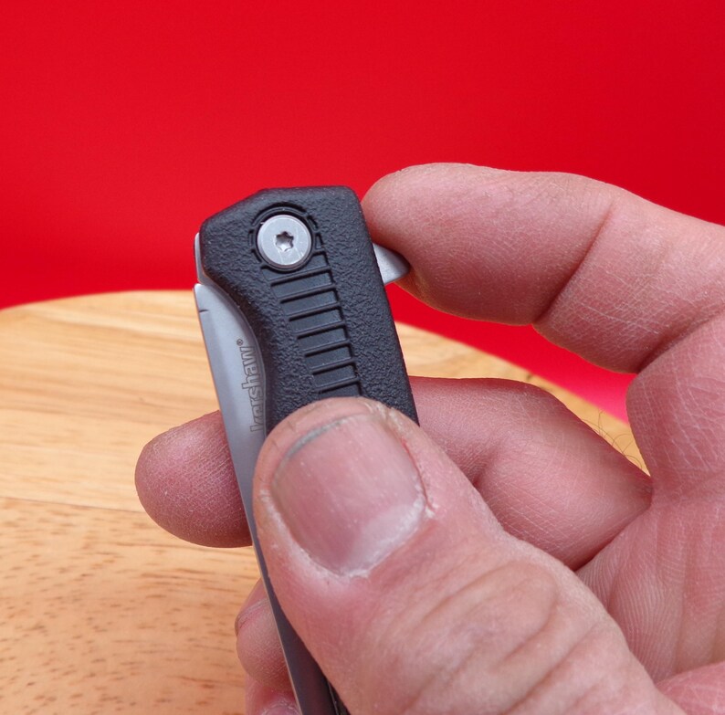 May include: A black and silver folding knife with a textured handle. The blade is partially open, revealing the silver metal. The knife is held against a red background.
