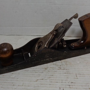 May include: A vintage wooden handled hand plane with a black metal body and a metal blade. The plane has a wooden knob on the front and a wooden handle on the back.