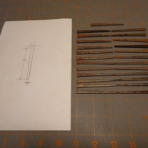 May include: A sheet of paper with a pencil drawing of a rectangle with measurements of 2 1/2 inches by 1/2 inch.  There are 20 rusty nails laid out in rows on a brown grid surface.