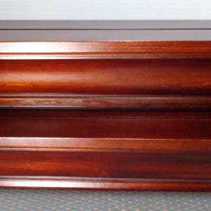 May include: Two-tiered, dark cherry-stained wooden shelf. The shelves have a classic design with a decorative, curved edge. The top shelf has a groove running along the top. The shelves are stacked on top of each other.