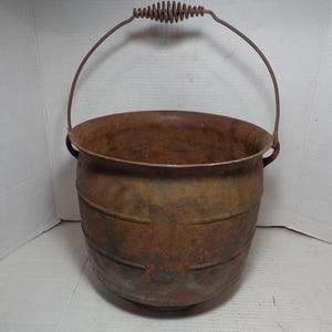 May include: A rusty cast iron bucket with a wire handle and a spring-loaded handle. The bucket has a rounded bottom and a wide opening.