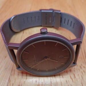 May include: A dark brown wristwatch with a matching mesh band. The watch face is dark brown with silver hands and markers. The watch has a date display at the 3 o'clock position. The watch is made of stainless steel.