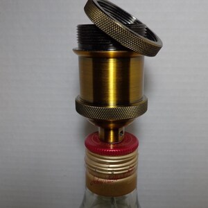 K&ouml;nnte beinhalten: Eine messingfarbene Lampenfassung mit strukturiertem Ring und schwarzem Innenraum, teilweise montiert. Die Fassung ist an einem Flaschenverschluss mit roten und goldenen Akzenten befestigt. Die Flasche ist aus klarem Glas.