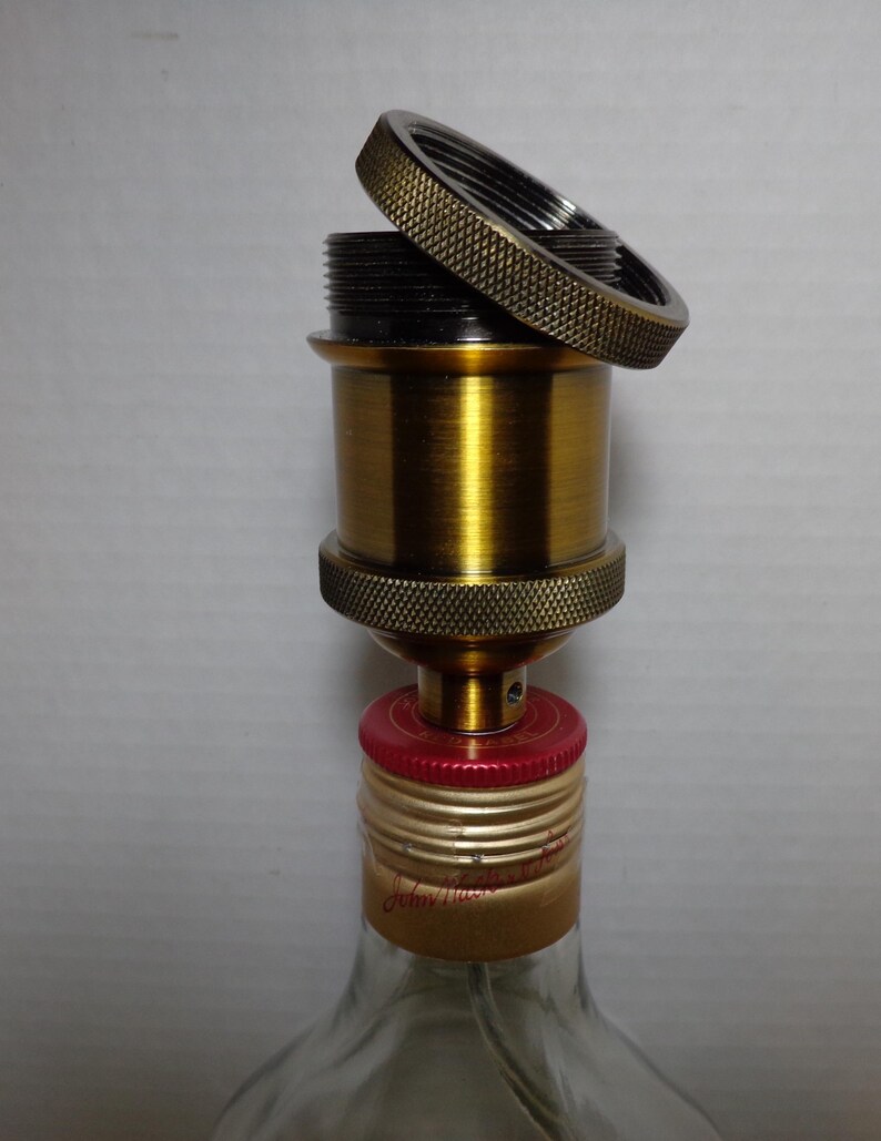 K&ouml;nnte beinhalten: Eine messingfarbene Lampenfassung mit strukturiertem Ring und schwarzem Innenraum, teilweise montiert. Die Fassung ist an einem Flaschenverschluss mit roten und goldenen Akzenten befestigt. Die Flasche ist aus klarem Glas.