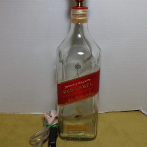 K&ouml;nnte beinhalten: Eine umfunktionierte Lampe aus einer Johnnie Walker Red Label Whiskyflasche. Die transparente Glasflasche hat ein rotes Etikett mit wei&szlig;em Text. Oben ist eine goldfarbene Lampenfassung angebracht, mit Netzkabel und Stecker.