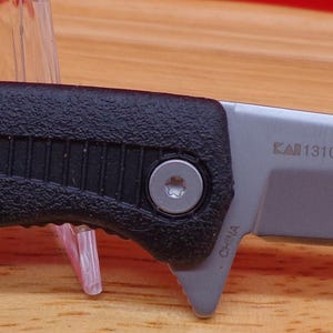 May include: A black and silver folding knife with a textured handle and a silver pocket clip. The blade is silver and has text on it. The knife is displayed on a wooden surface.