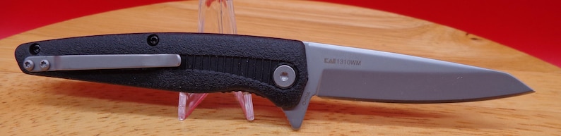 May include: A black and silver folding knife with a textured handle and a silver pocket clip. The blade is silver and has text on it. The knife is displayed on a wooden surface.