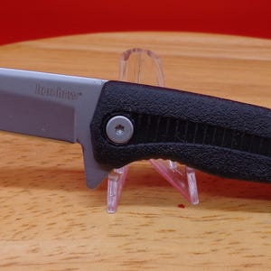 May include: A black and silver folding knife with a textured handle. The blade is silver and angled, and the handle has a ridged design. The knife is displayed on a wooden surface.