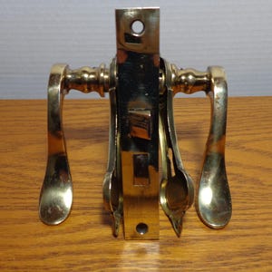 May include: A vintage brass door handle set with a rectangular backplate and curved lever handles. The set includes a latch mechanism and is designed for a door. The brass has a warm, aged patina.