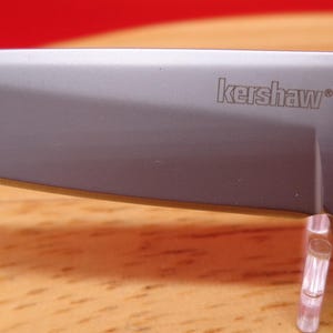 May include: Close-up of a silver folding knife blade with the brand name "kershaw" etched on the blade. The knife has a black handle and a silver blade. The knife is on a wooden surface.