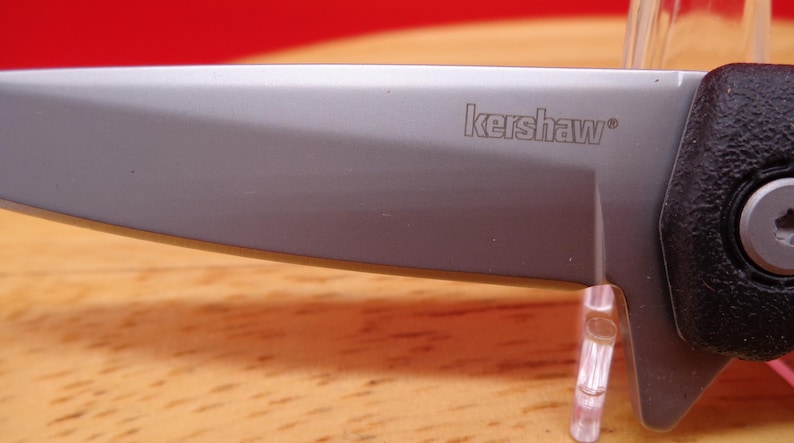 May include: Close-up of a silver folding knife blade with the brand name "kershaw" etched on the blade. The knife has a black handle and a silver blade. The knife is on a wooden surface.