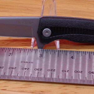 May include: A black and silver folding knife with a partially serrated blade. The knife is shown with a ruler, and the blade appears to be approximately 3 inches long. The handle is black and textured.